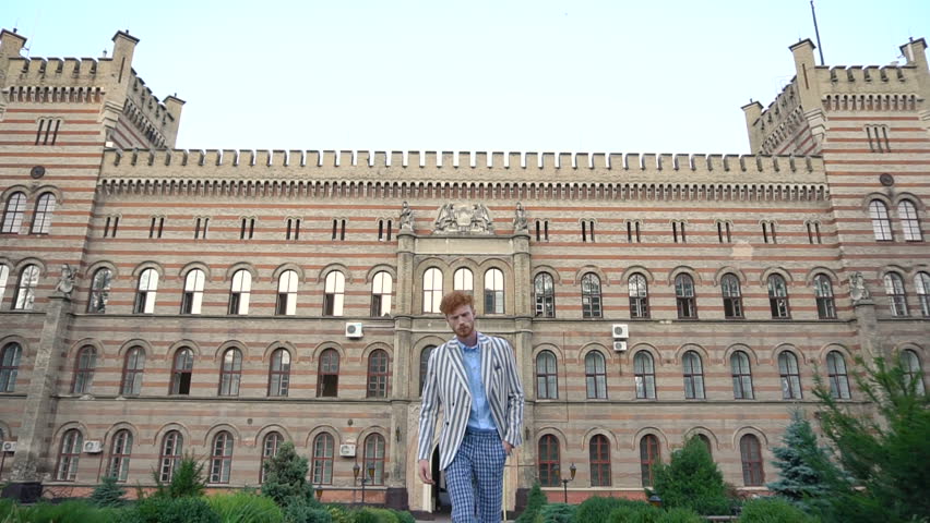 Confident attractive man with curly ginger hair in trews pants and striped jacket walks at the background of the old building. He stops, looks away and goes back.