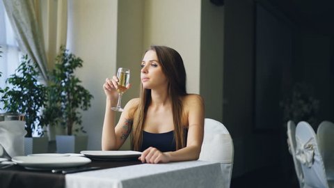 Pretty young woman is sitting alone at table in restaurant, drinking champagne and waiting for her boyfriend. Girl is nervous and feeling uncomfortable.