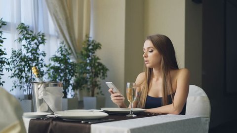 Angry beautiful woman is calling her boyfriend on mobile phone while waiting for him alone in restaurant, then leaving. Girl is impatient, nervous and offended.