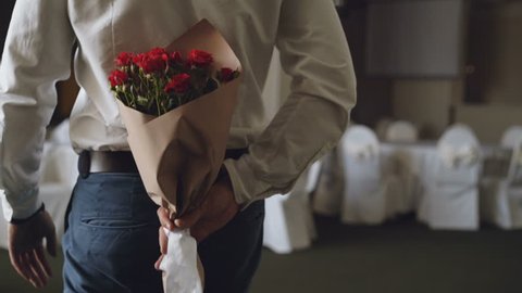 Loving man is hiding red roses behind his back bringing beautiful bouquet for his date in restaurant. Flowers, romantic relationship and dating concept.