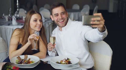 Attractive loving couple is taking selfie with champagne glasses using smartphone while having dinner in restaurant. They are smiling, kissing and posing looking at camera.
