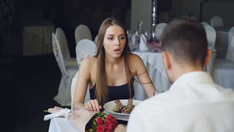 Angry young woman is quarreling with her lover while dining in restaurant, shouting and gesturing. Bouquet of flowers and plate with food are visible.