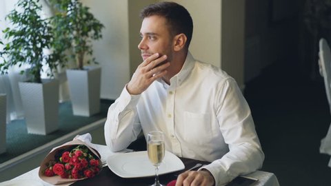 Nervous man is sitting alone at table in restaurant, drinking champagne and waiting for his girlfriend, then leaving. Bunch of red roses, jewelry box and smartphone are visible.