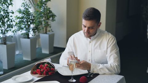 Well-dressed angry young man is waiting for his girlfriend in restaurant, using smartphone, opening jewelry box and looking at ring then leaving, flowers left on table.
