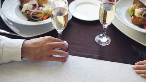 Close-up shot of young lovers touching and holding hands in classy restaurant. Table with sparkling champagne glasses, flatware and food in background.