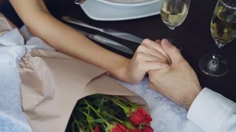 Close-up shot of male hand holding and squeezing female hand on table with champagne glasses and flowers. Romantic relationship, love and fine dining concept.
