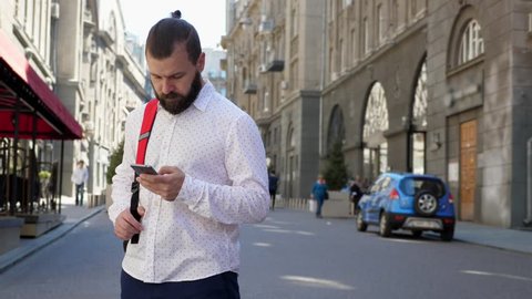 A bearded man on a European street reads messages in his smartphone and looks around.