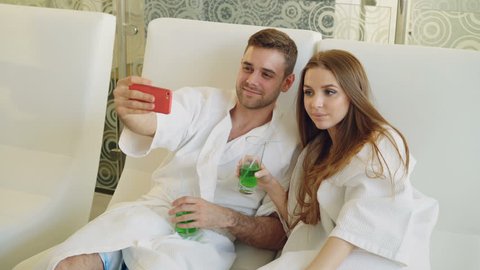 Attractive loving couple is taking selfie with cocktail glasses using smartphone while relaxing in spa salon. They are smiling and posing looking at camera.