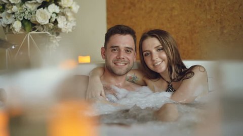 Portrait of good-looking young lovers hugging in jacuzzi, smiling and looking at camera. Burning candles, beautiful flowers and bubbling water is visible.