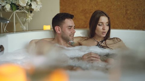 Adorable young couple is having fun in bubbling hot tub playing with foam, talking and kissing. Romantic relationships, happy people and wellness concept.