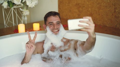 Handsome guy is taking selfie in bathtub with soap foam on his beard using smart phone. He is laughing and making gestures with his hand posing and having fun.