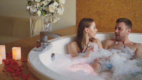 Attractive cheerful couple in hot tub is playing blowing foam, kissing and enjoying romantic moments. Rose petals, flowers and burning candles are visible.
