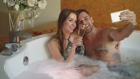 Pretty girl and her loving boyfriend are taking selfie with champagne glasses using smartphone while bathing in bathtub. They are smiling and posing looking at camera.