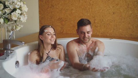 Cheerful pretty girl in bathing suit and her handsome boyfriend are playing with soap foam in jacuzzi in spa salon. Happy people, relaxation and wellness concept.