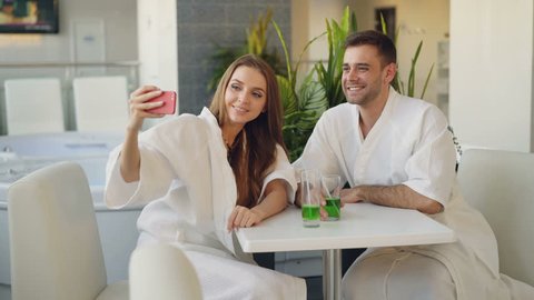 Pretty girl and her loving boyfriend are taking selfie with cocktail glasses using smartphone while relaxing in spa salon. They are smiling and posing looking at camera.