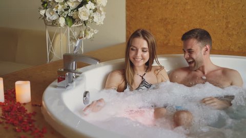 Pretty cheerful girl is blowing soap foam and having fun with her bearded fience in hot tub in day spa. Young people are laughing, enjoying weekend and talking.