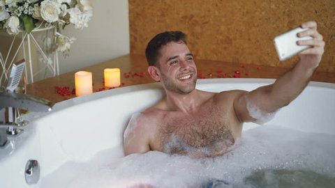 Young bearded guy famous blogger is recording video in hot tub in day spa using smartphone. Burning candles, champagne glass and flowers are visible.