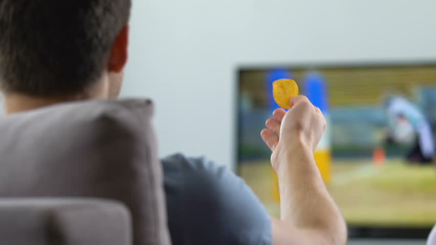 Emotional football fan watching match on TV at home, eating chips, back view