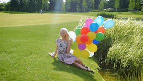 Slow motion video of happy young woman in dress sitting on grass with balloons