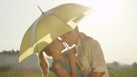 SLOW MOTION, CLOSE UP, LENS FLARE: Cute young couple smiles and kisses in the refreshing summer rain. Cheerful man dips and kisses his smiling girlfriend while they dance in the quiet countryside.