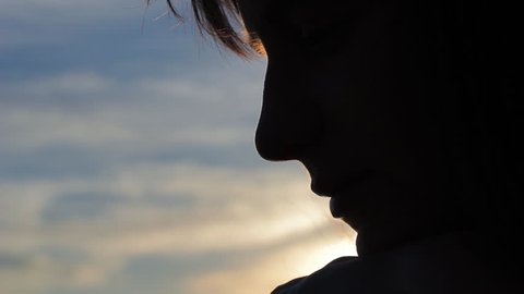 Sad thoughtful woman sitting in solitude against sunset sky. Close up profile silhouette of calm young woman sitting with her head on her knees outdoor, panning shot