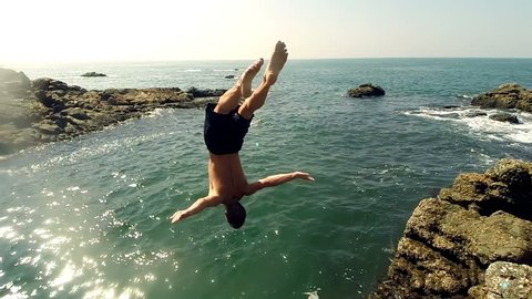 Male athlete jumping from a cliff into the water. Athletic young man doing back flips into sea. Slow-motion.