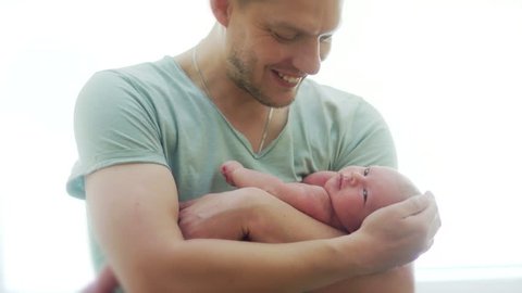 A young father of athletic builds a naked newborn baby on his hands. A man strokes him on the head, the child waves with a pen and turns his head, looks around. Father's Day