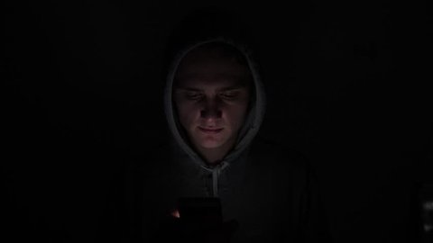 Hooded teen with a dark black background in shadow using a mobile swiping device with thumbs, close up shot.