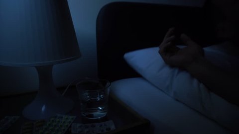 Man sleeping bad in bed at night home. Suffering from insomnia sickness headache. Medications pills tablets, glass of water, night lamp on bedside table. Self-medication concept. Filmed 4K Sony a6300.