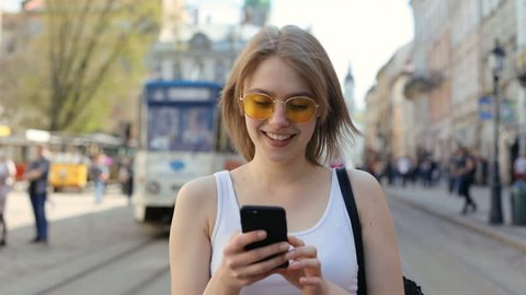 portrait of walking relaxed smiling girl using digital smartphone mobile outdoors blurred tramway on background internet connecting city space 3G traffic app comfortable lifestyle casual clothes