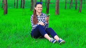 Female person listen to music by phone in ear phones, sitting on grass. Concept of speaking by modern gadgets and fast Internet in park