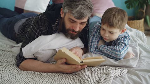 Loving parent bearded man is reading interesting book to his curious little son on double bed at home. Parenthood, happy people, love and family concept.