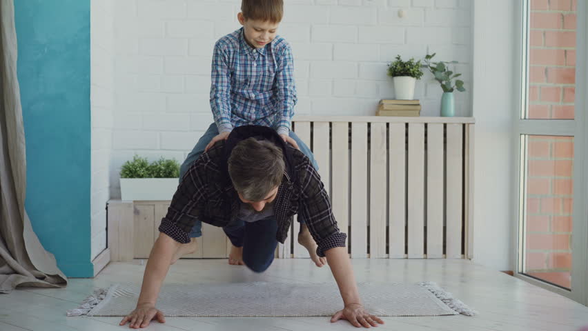 Strong active man is doing push-ups with his laughing little son sitting on his back. Sport, spending time with family, happy childhood concept.