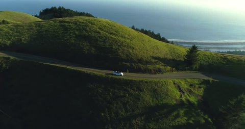 Aerial view of car driving down country road in rural rolling hills with ocean in background at sunset