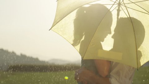 SLOW MOTION, CLOSE UP, LENS FLARE: Couple hidden behind a yellow umbrella kisses while dancing in the spring rain. Carefree Caucasian man and woman on a fun date in golden lit nature spin and kiss.