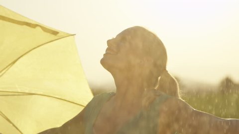 SLOW MOTION, CLOSE UP: Carefree young blonde woman lets the refreshing spring rain fall down on her. Cheerful girl in green sundress puts her umbrella to the side and dances in the cool summer rain.