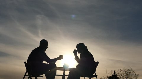 2 men silhouettes drink tea and communicate outdoors at sunset