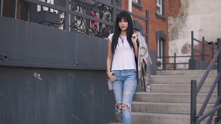 the fashionable girl goes down the stairs. Portrait of an attractive girl on a background of architecture. a young woman in ripped jeans and a T-shirt is throwing her jacket over her shoulder.