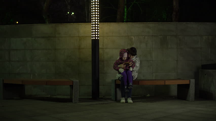 Mother and daughter sit on a bench under the light of a lantern and use the phone at night in the park. A woman is reading to a child.