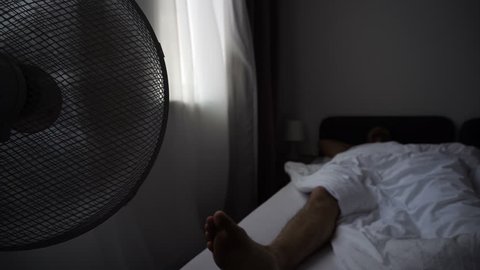 Electric fan turning around, blowing fresh cold air, moving curtain on windows. Man resting or sleeping in hotel room bed this warm hot morning. Cooler for summer weather promotion concept.