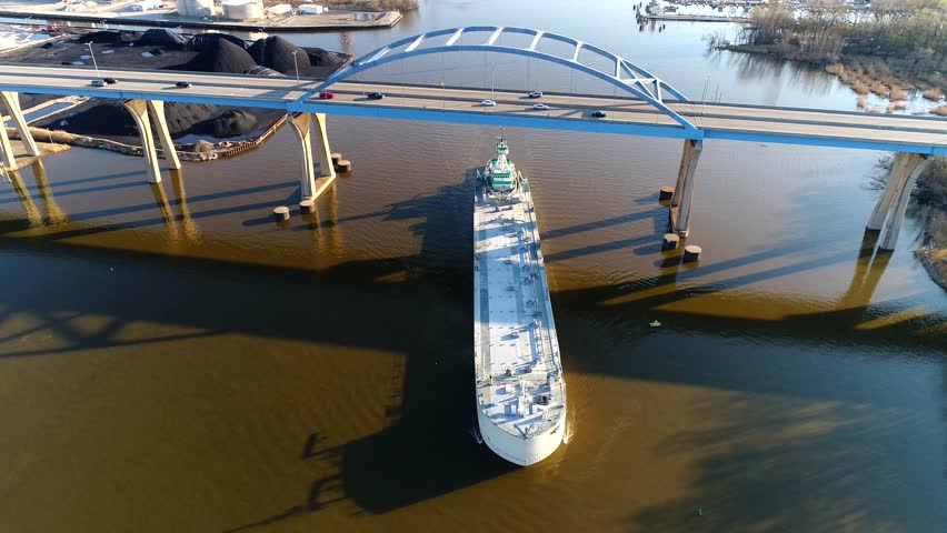 Freighter ship sails under busy bridge, early morning aerial view.
