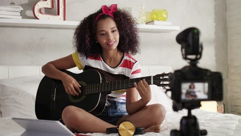 Happy black girl at home playing guitar. Young African American woman working as web influencer, recording vlog and music tutorial for the Internet.