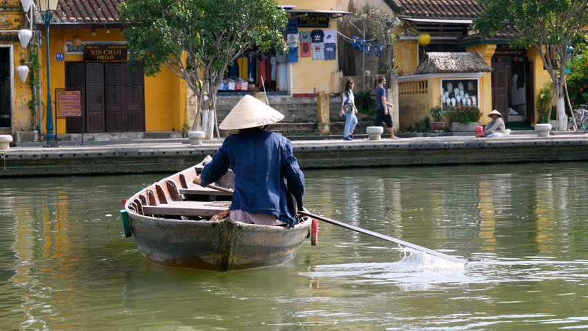 Static rear shot of a solo Vietnamese boat taxi woman paddling across a small river in Hoi An to pick up a waiting pedestrian, both wearing non la hats.