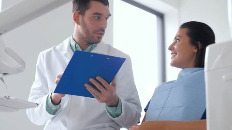 medicine, dentistry and healthcare concept - male dentist with clipboard talking to female patient at dental clinic office