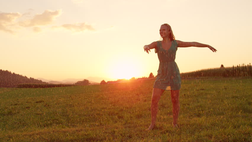 SLOW MOTION, LENS FLARE, COPY SPACE: Pretty Caucasian woman twirls elegantly in a cool spring shower at sunset. Stunning shot of happy blonde girl dancing in the morning in beautiful summer nature. | Shutterstock HD Video #1010844662