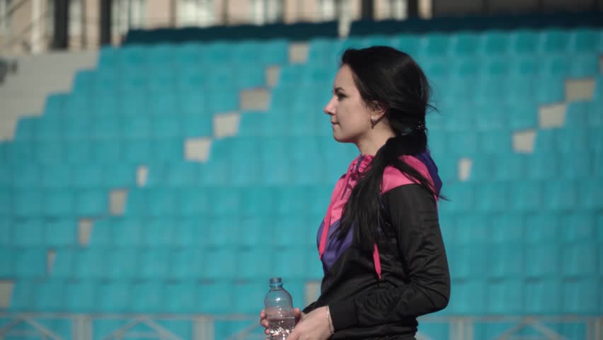 Sexy slim athletic woman drinks water after practicing on a stadium background