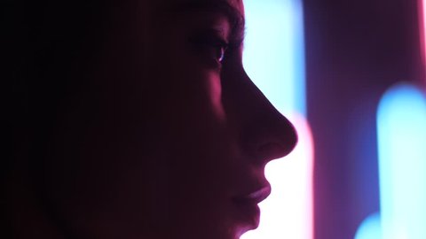 closeup side view beautiful young woman blinks and looks sadly forward against pink blue night light – Video có sẵn