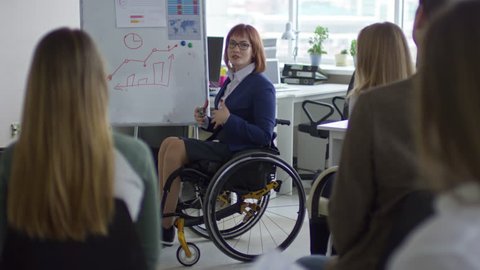 Tilt up of female business coach sitting in wheelchair and explaining bar graph on flipchart to group of office workers