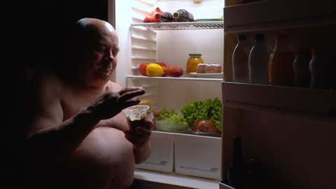 A fat man sits next to the refrigerator at night and eats chocolate mousse.