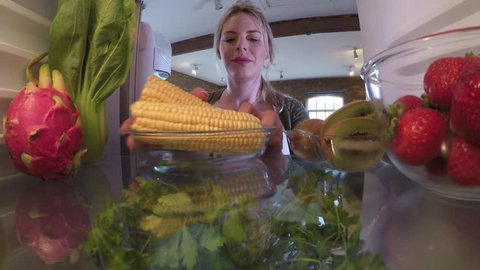 A POV shot from inside a fridge showing a woman opening the fridge to take out some corn.